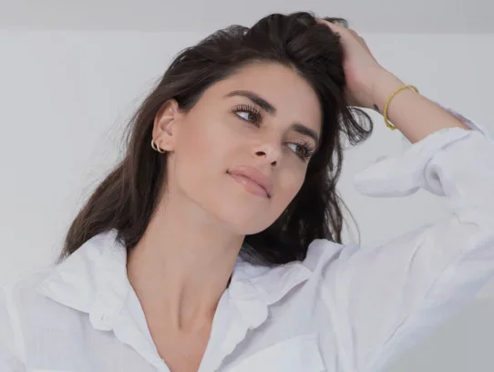 Woman in white shirt with hand in dark hair looking confidently at camera against light background - Dermal fillers in Evansville, IN