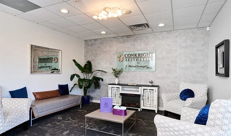 Modern office waiting room with white furniture, fireplace, potted plant, and contemporary artwork