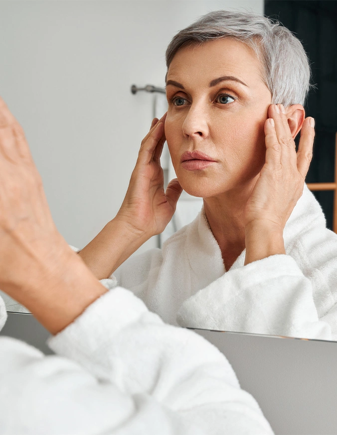 Woman with short blonde hair examining her face in mirror checking for signs of aging and wrinkles - Eyelid surgery in Evansville, IN