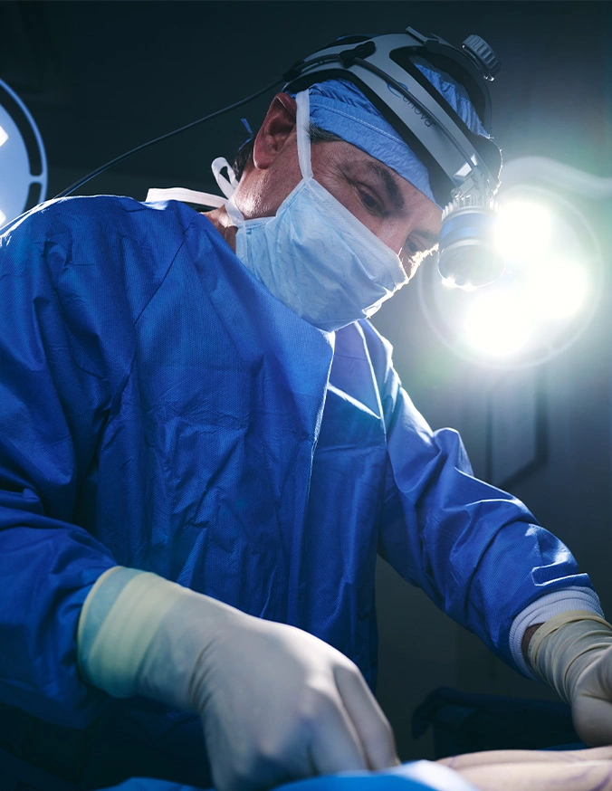 James Micheal Conkright MD in blue scrubs and protective gear performing operation under surgical lights in operating room - Liposuction in Evansville, IN