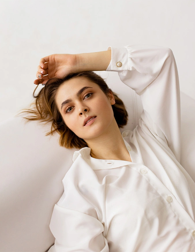 Woman in white shirt with tousled auburn hair posing with arm raised against neutral background - Brow lift in Evansville, IN