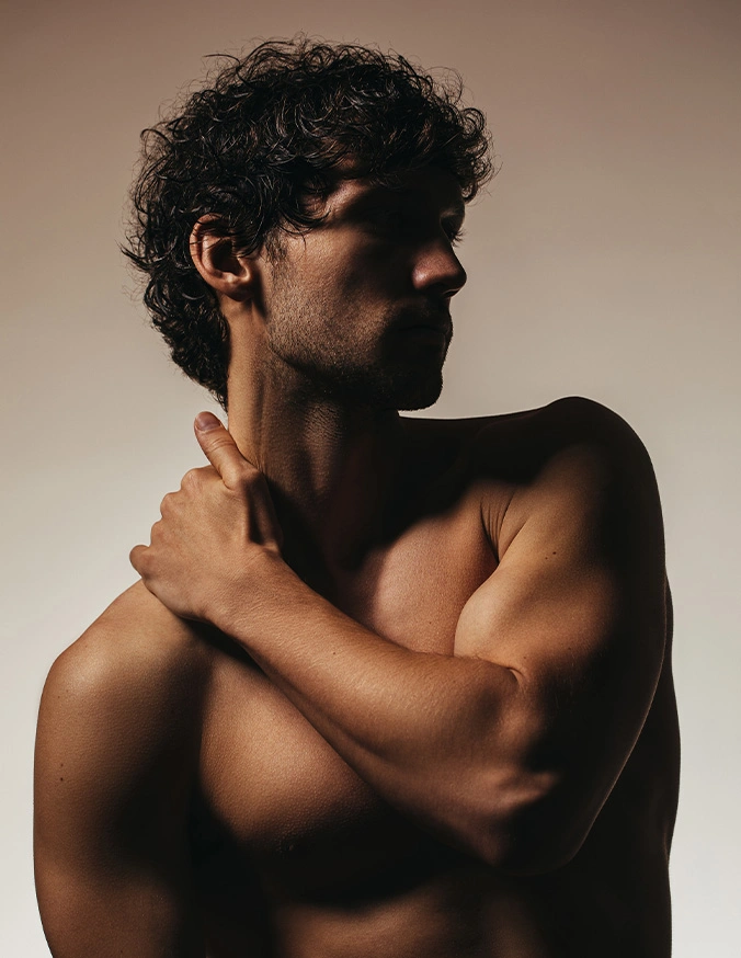 Shirtless man with curly hair in profile view embracing himself against neutral background - Gynecomastia in Evansville, IN